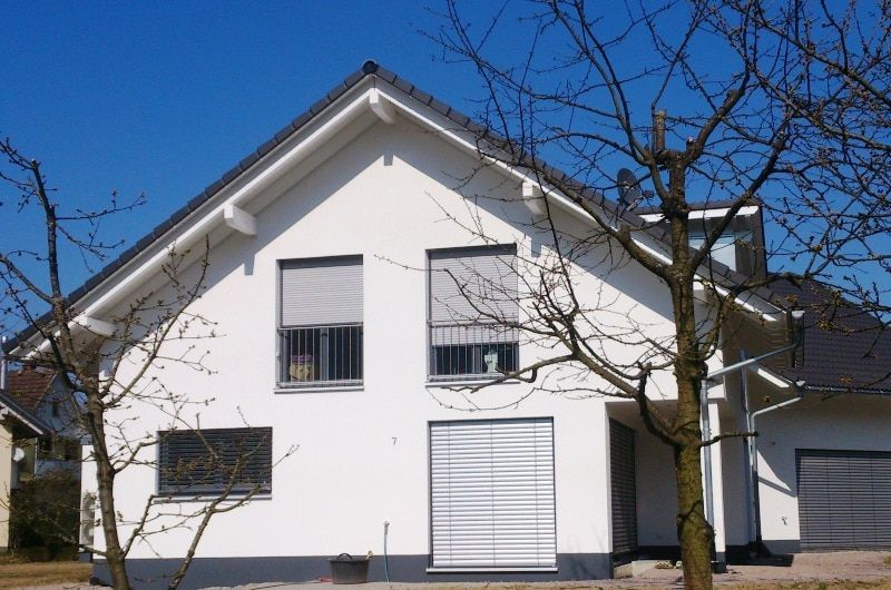 Weißes Haus mit dunklem Dach und grauen Jalousien, umrahmt von kahlen Bäumen unter blauem Himmel.