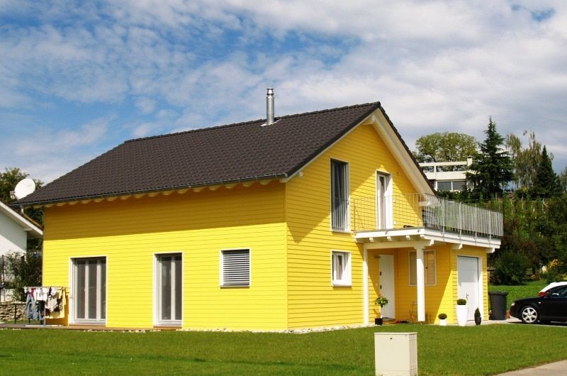 Gelbes Haus mit schwarzem Dach und weißen Zierelementen unter blauem Himmel.