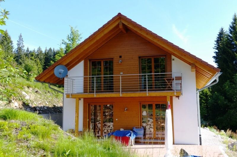 Zweistöckiges Haus mit Holzverkleidung, Balkon und Satellitenschüssel. Weiße Fassade, am Hang gelegen.