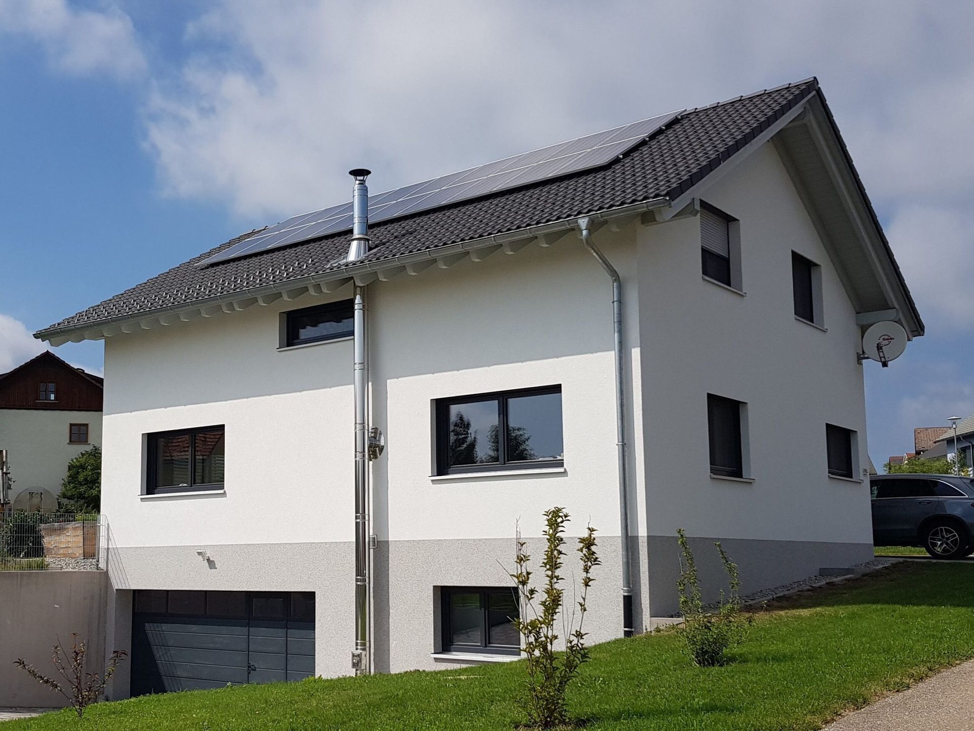 Zweistöckiges weißes Haus mit dunklem Dach und Solarpaneelen, grauem Untergeschoss und grünem Rasen.