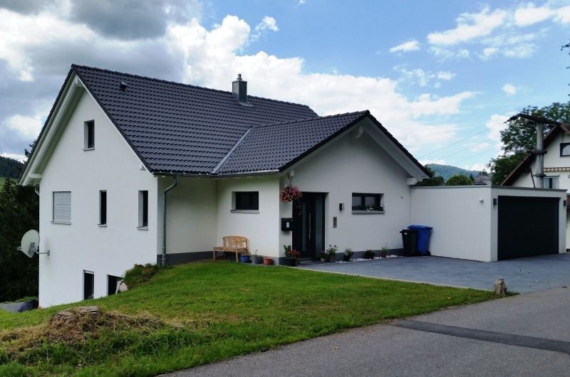 Weißes Haus mit dunklem Dach und angebauter Garage, auf einem Hügel mit grünem Rasen und Auffahrt, unter bewölktem Himmel.