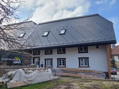 Weißes Haus mit dunkelgrauem Dach, mehreren Fenstern und Baumaterialien.