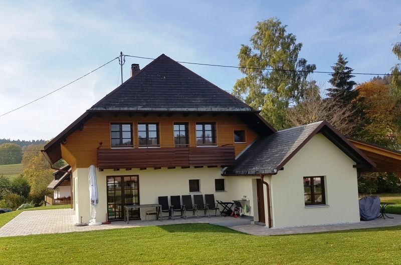 Haus mit braunem Dach und Wänden, hellbraunem Untergeschoss, Balkon und kleinem Schuppen, in einem grasbewachsenen Hof.