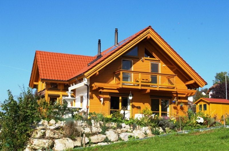 Holzhaus mit orangefarbenem Ziegeldach, Balkon und steinerner Stützmauer unter blauem Himmel.