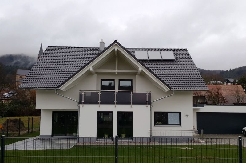 Weißes Haus mit dunkelgrauem Dach, Balkon und Solaranlage. Schwarze Fensterrahmen und Garage.