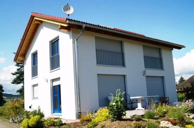 Zweistöckiges weißes Haus mit blauen Türen und Fensterläden, einer Satellitenschüssel und einem Garten vor blauem Himmel.