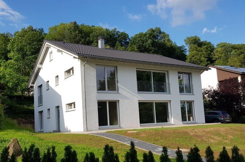 Zweistöckiges weißes Haus mit großen Fenstern, gelegen auf einem grünen Rasen vor einer Kulisse aus bewaldeten Hügeln und blauem Himmel.