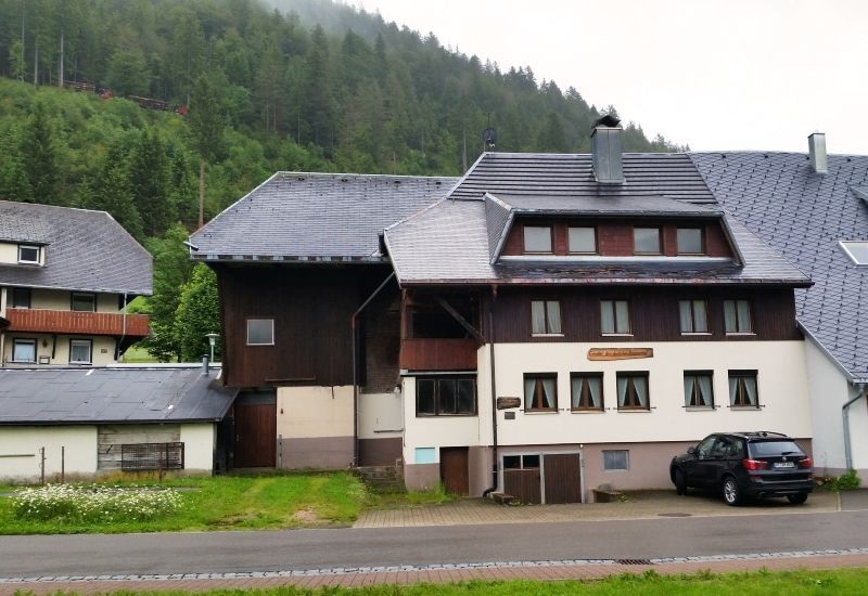 Gebäude mit dunklem Dach und braun-weißer Fassade, geparktes Auto, grüner Hügel im Hintergrund.