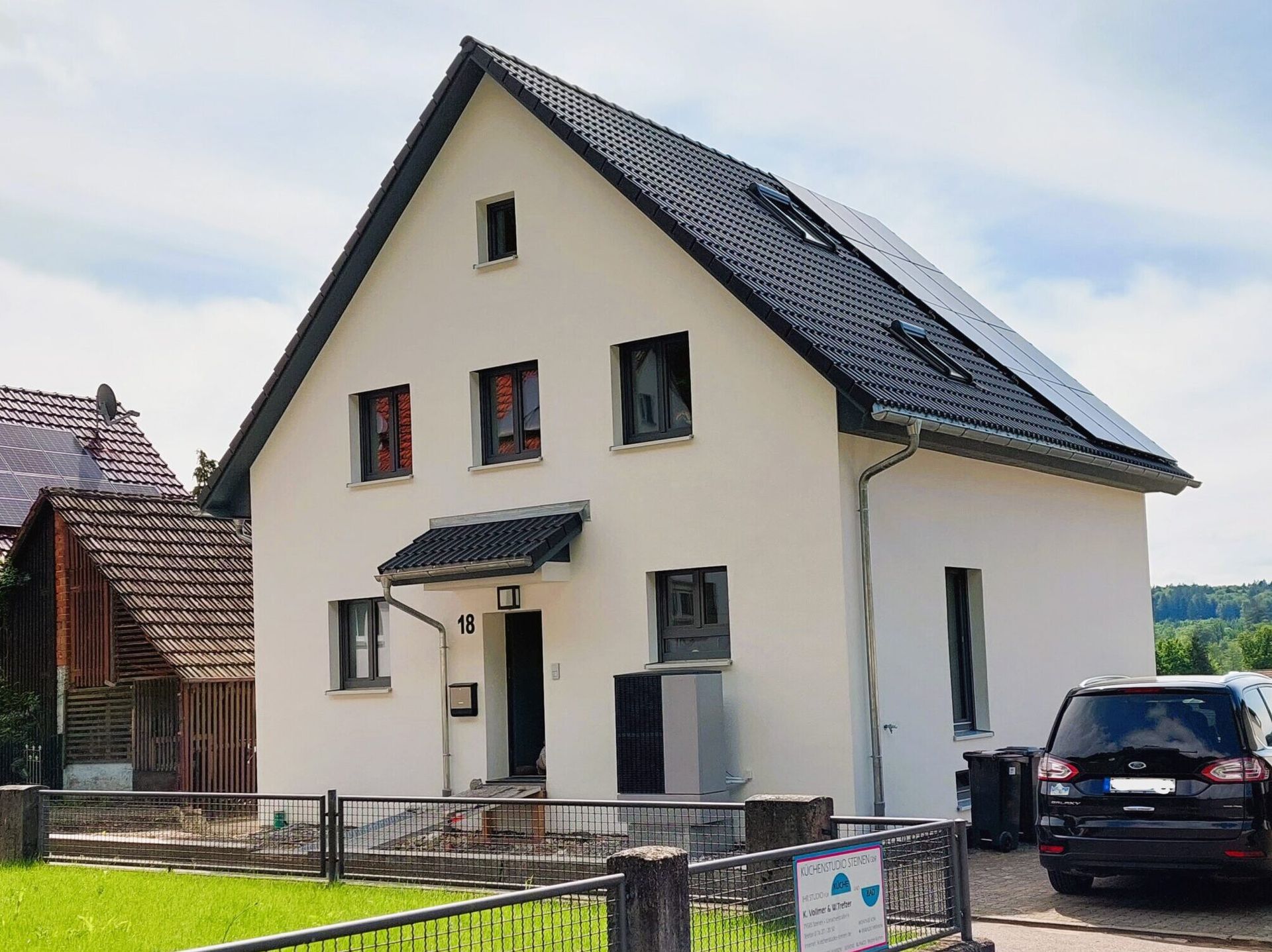 Zweistöckiges Haus mit schwarzem Dach und Solarpaneelen. Weiße Wände, schwarze Tür und ein geparkter schwarzer Geländewagen.