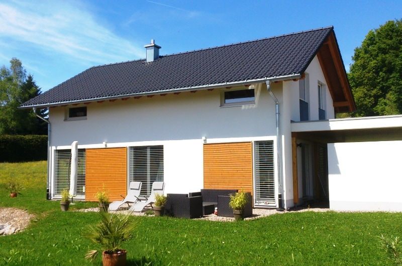 Modernes weißes Haus mit schwarzem Dach und Holzfensterläden, sonniger Tag.