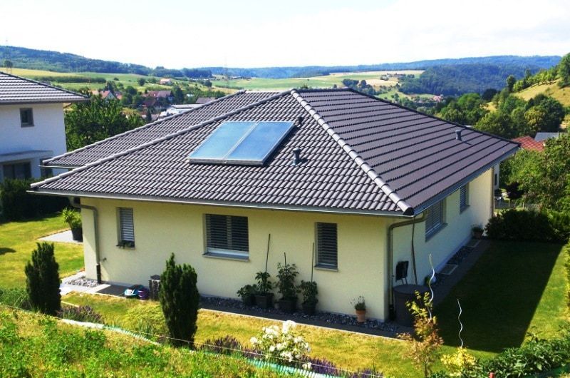 Beiges Haus mit dunklem Dach und Solaranlage, eingebettet in eine grüne Landschaft mit sanften Hügeln.