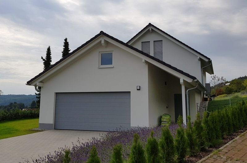 Ein zweistöckiges weißes Haus mit grauem Garagentor und Dach, Einfahrt und Gartengestaltung.