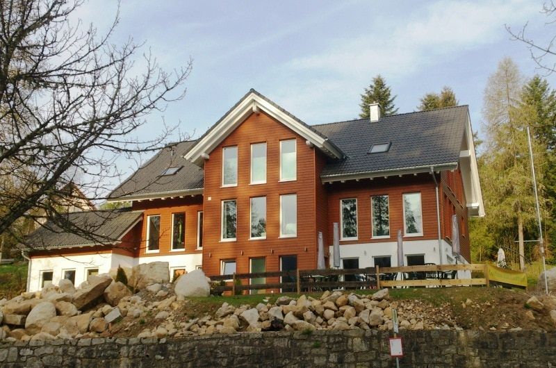 Backsteingebäude mit vielen Fenstern und dunklem Dach, auf einem Hügel gelegen, im Vordergrund Felsen.