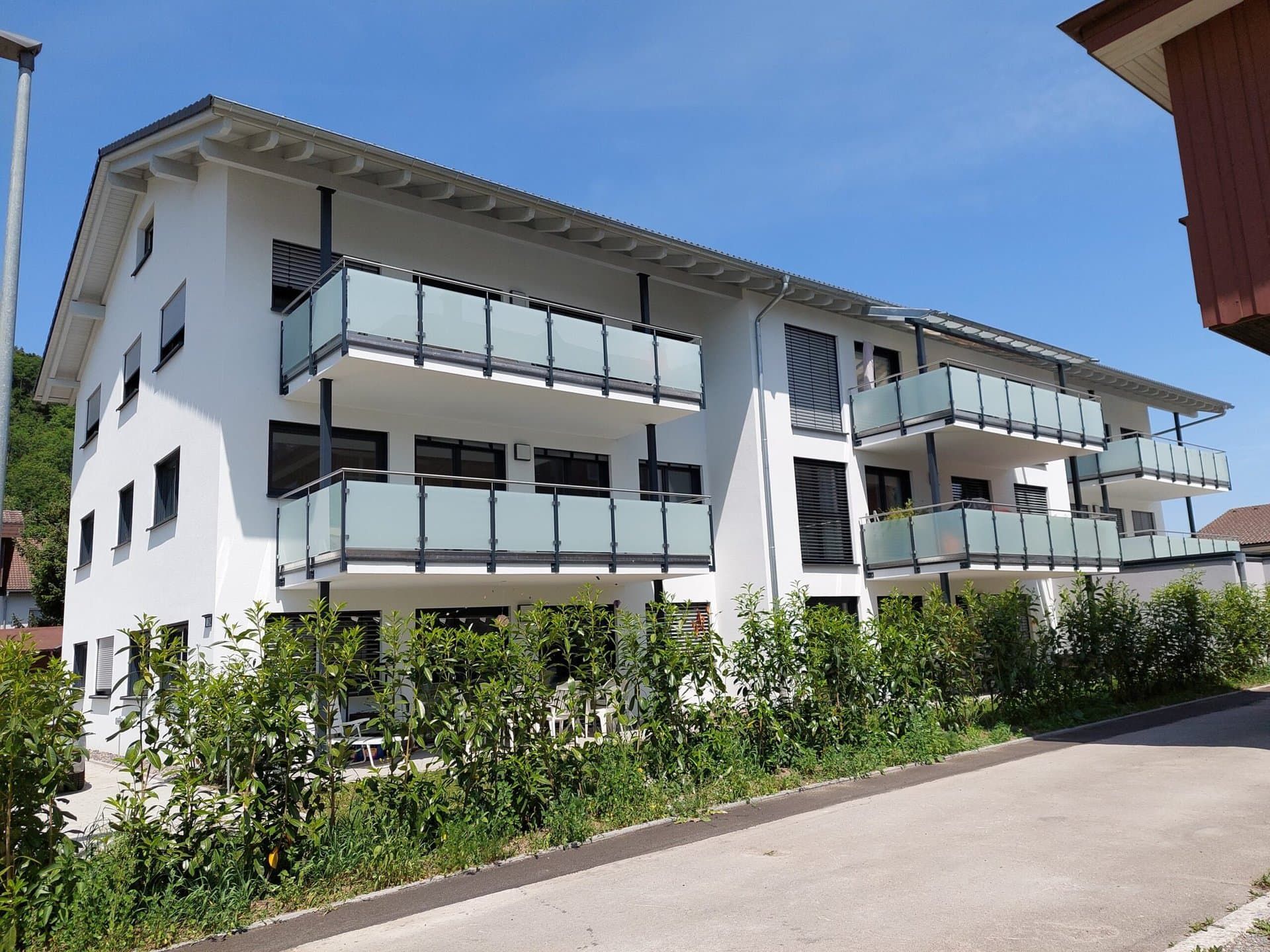 Weißes Wohnhaus mit Balkonen, Sträuchern und einer Straße an einem sonnigen Tag.