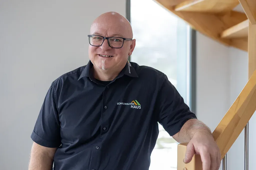 Ein Mann mit Brille und Glatze, in einem dunklen Hemd, lächelt neben einer Holztreppe.