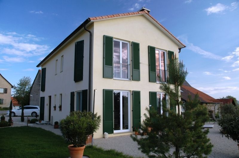 Zweistöckiges beiges Haus mit grünen Fensterläden vor blauem Himmel, in dessen Einfahrt ein Auto steht.