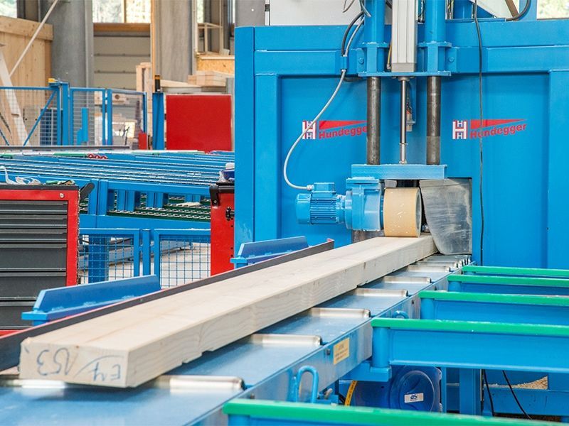 Eine blaue Industriemaschine schleift einen langen Holzbalken auf einem Förderband.