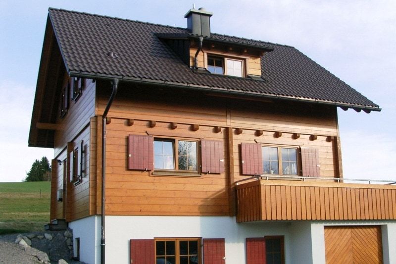 Zweistöckiges Holzhaus mit braunem Dach und roten Fensterläden, gelegen auf einem grasbewachsenen Hügel.