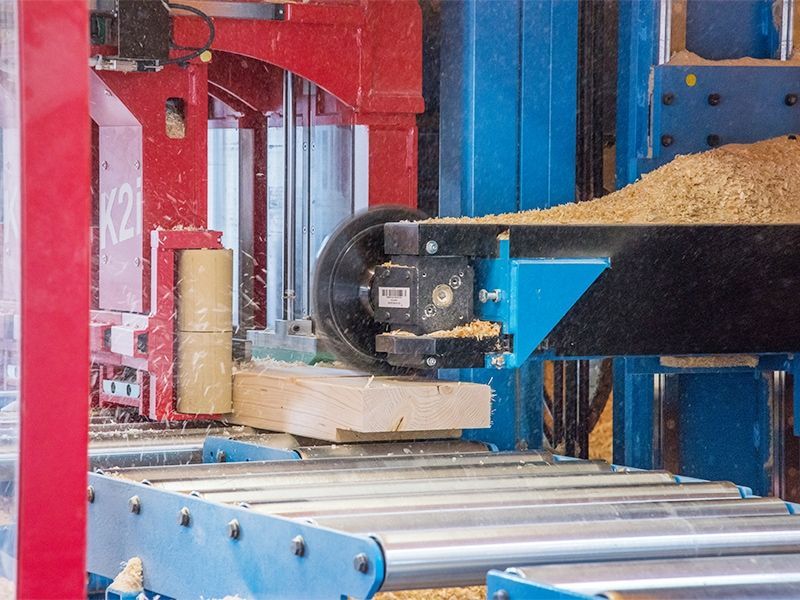 Eine große Industriesäge schneidet Holz auf einem Förderband; Sägemehl und Maschinenteile sind zu sehen.