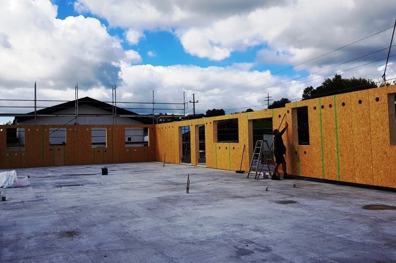 Baustelle mit im Bau befindlichen Holzwänden, eine Person arbeitet auf einer Leiter. Betonboden, bewölkter Himmel.