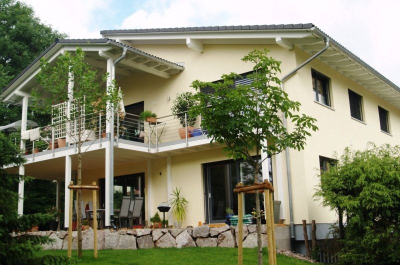 Zweistöckiges beigefarbenes Haus mit Balkon und Rasen. Bäume im Garten, Steinmauer am Fuße des Hauses.