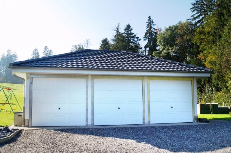 Dreifachgarage mit weißen Toren und grauem Ziegeldach, gelegen an einer Kiesauffahrt, im Hintergrund Bäume.