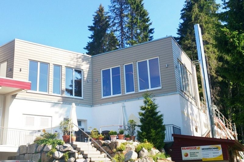 Modernes Gebäude mit großen Fenstern und einem Schornstein, umgeben von Bäumen.