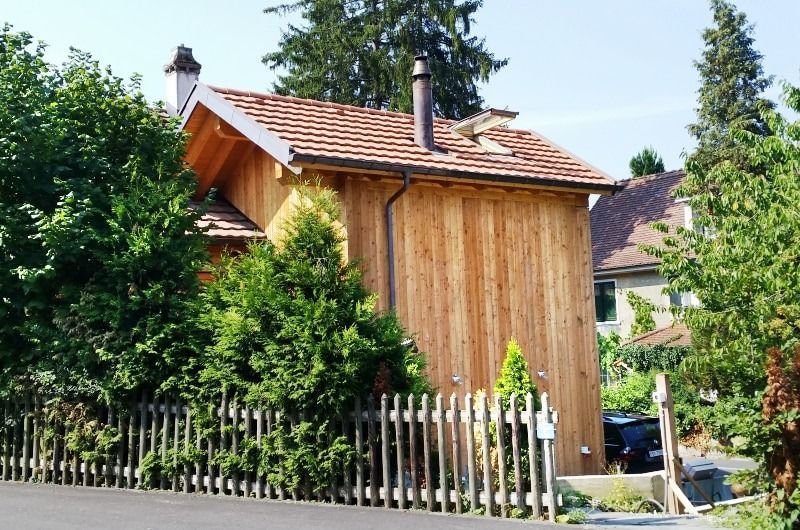 Holzhaus mit braunem Ziegeldach und Schornstein, hinter einem Holzzaun und Bäumen.