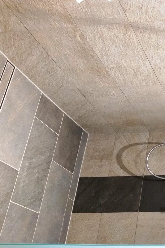 Badezimmerecke mit grauen und beigen Fliesen, Abfluss und einem silbernen Handtuchring.