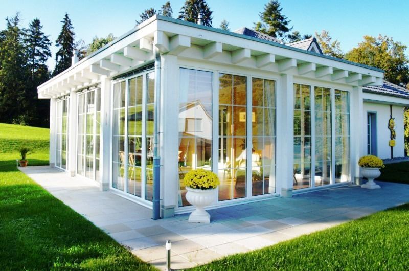 Weißer Wintergarten mit großen Fenstern, Steinterrasse und gelben Blumentöpfen. Gelegen auf einem grünen Rasen mit Bäumen im Hintergrund.