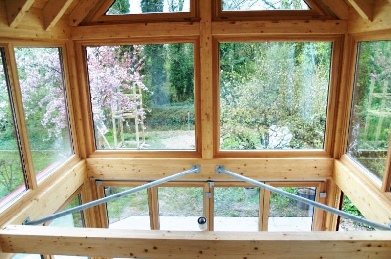 Innenraum eines hölzernen Pavillons mit großen Fenstern und Blick auf einen Garten mit Bäumen und Grünanlagen.