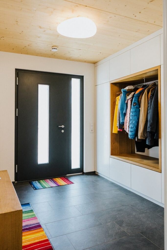 Eingangsbereich mit schwarzer Tür, Garderobe und farbenfrohem Teppich. Weiße Einbauschränke und Holzdecke.