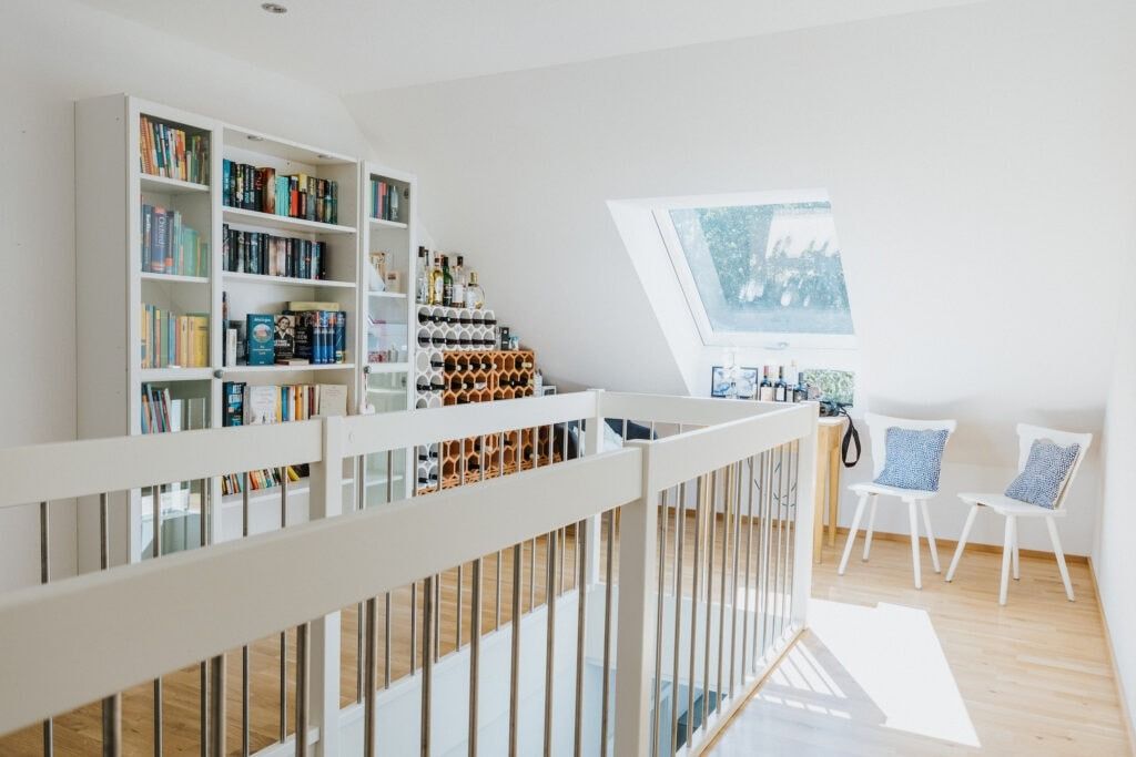 Heller, luftiger Dachbodenraum mit Bücherregalen, einem Weinregal, einem Geländer und zwei weißen Stühlen am Fenster.