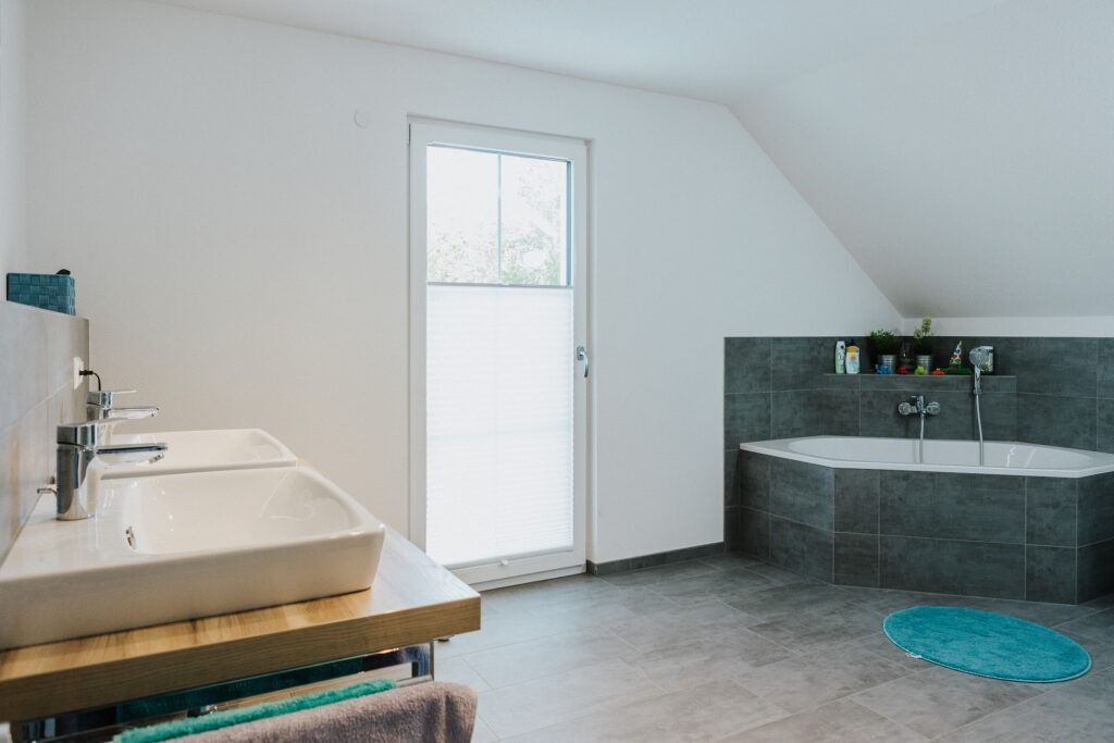 Badezimmer mit zwei Waschbecken, einer Badewanne und einer Tür, mit grauen Fliesen und einem hellblauen Teppich.