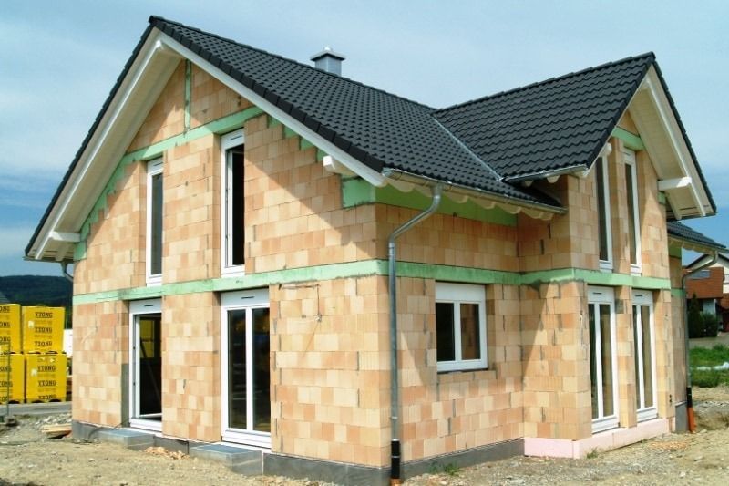 Haus im Bau, Außenansicht, mit ungestrichenen Ziegelwänden, weißen Fensterrahmen und schwarzem Dach.