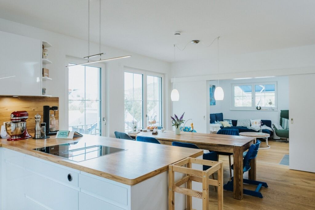 Helle Küche mit Kochinsel, Esstisch und Wohnbereich im Hintergrund. Weiße Wände, Holzböden, blaue Stühle.