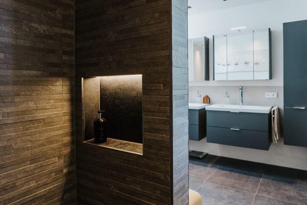 Modernes Badezimmer mit grau gefliester Dusche und eingelassenem Regal mit einer Flasche. Dunkle Schränke und weiße Arbeitsplatte sind sichtbar.