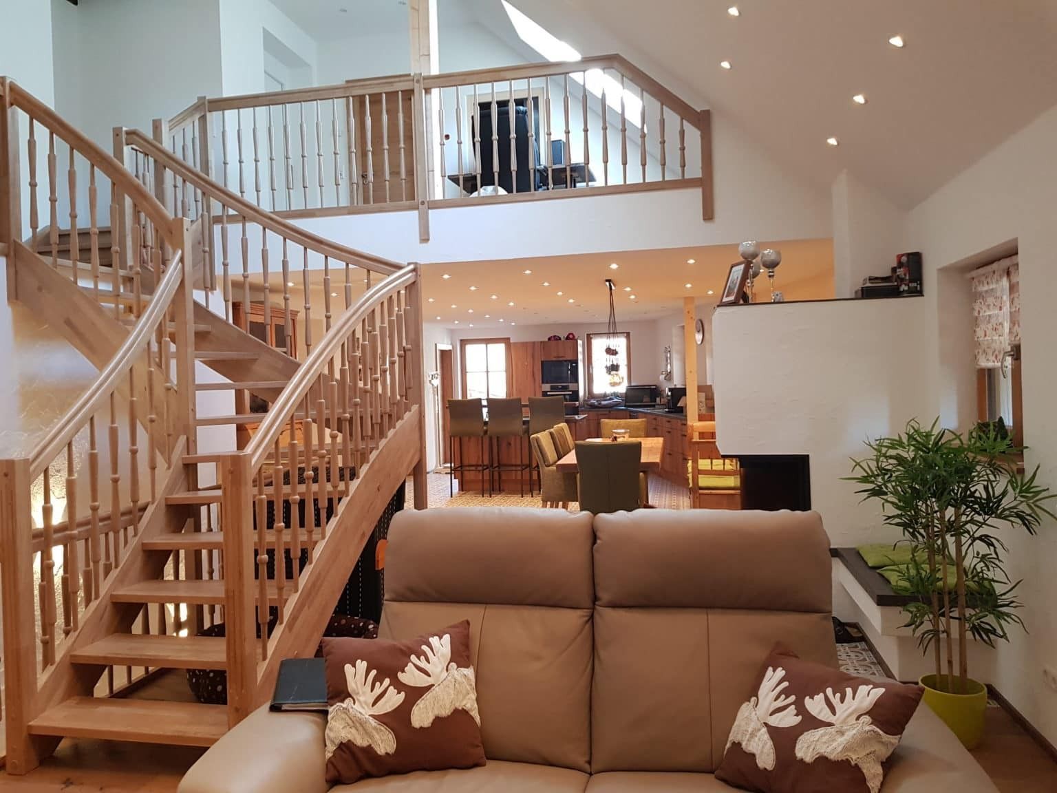 Eine Holztreppe führt zu einem Dachboden mit Blick auf einen Wohnbereich mit braunem Sofa und im Hintergrund eine Küche.