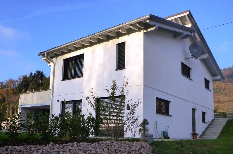Weißes zweistöckiges Haus mit schwarz umrandeten Fenstern, grauem Dach und Satellitenschüssel. Sonniger Tag.