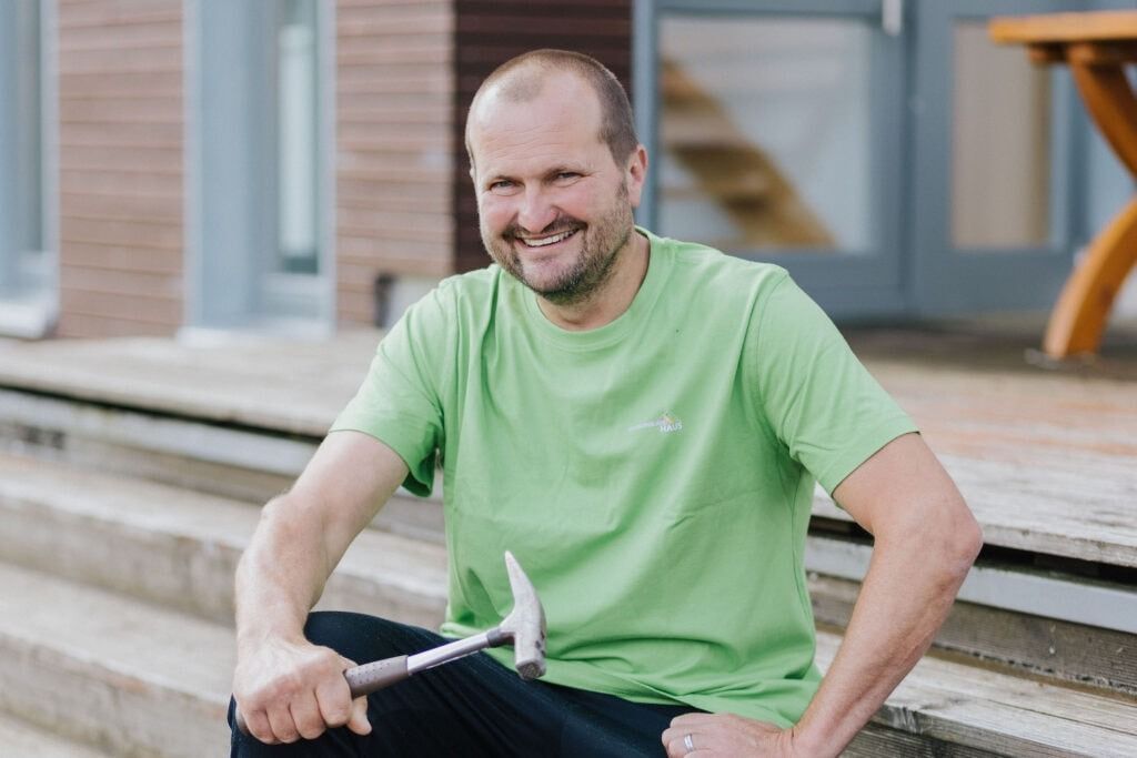 Ein Mann in grünem Hemd lächelt, hält einen Hammer in der Hand und sitzt auf einer Außentreppe.