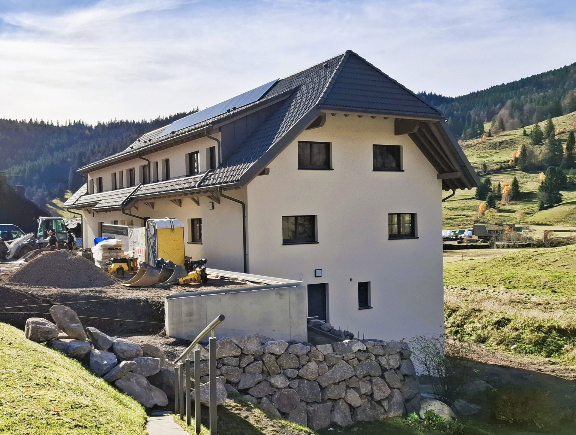Weißes Gebäude mit dunklem Dach, Solarpaneelen und Steinmauer auf einer Wiese. Baumaschinen in der Nähe.