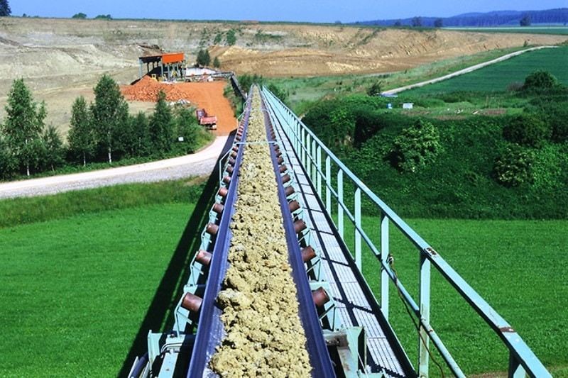 Förderband transportiert Material über ein grünes Feld, das zu einem Steinbruch und Gebäuden führt; sonniger Tag.