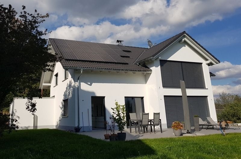 Weißes zweistöckiges Haus mit dunkelgrauem Dach und Fensterläden, Gartenmöbel auf einer Rasenfläche unter bewölktem Himmel.