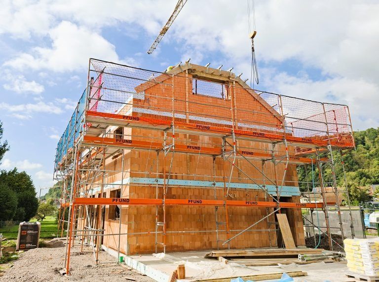 Baustelle mit Backsteingebäude, Gerüst, Sicherheitsnetz und Kran unter bewölktem Himmel.