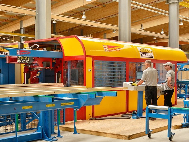 Zwei Männer bedienen in einer Fabrik eine große, gelb-rote, automatische Holzverarbeitungsmaschine.