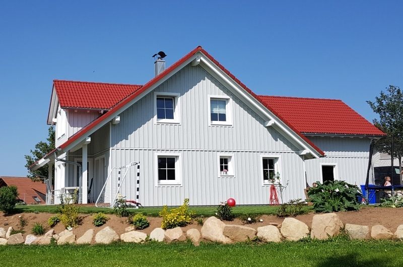 Weißes Haus mit rotem Dach unter blauem Himmel, mit einem kleinen Garten davor.