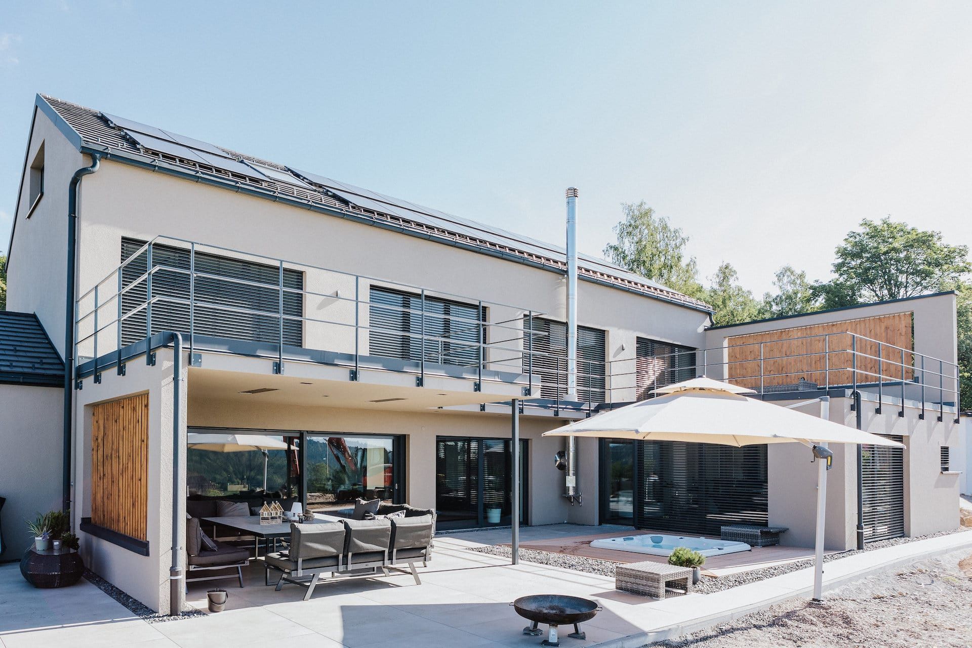 Modernes zweistöckiges Haus mit Terrasse, Balkon und Essbereich im Freien. Graue Fassade, Sonnenschirm, Feuerstelle und sonniger Tag.