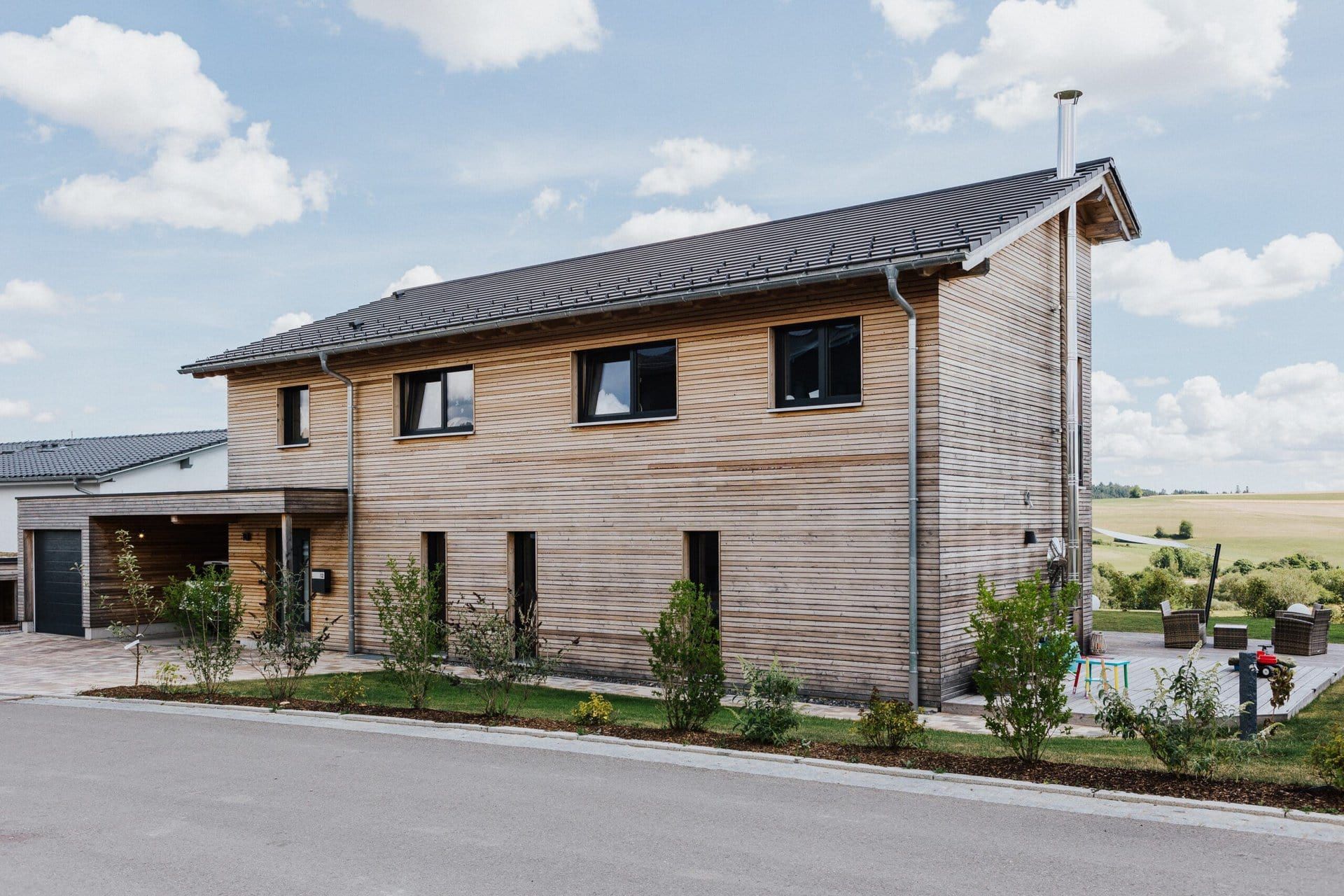 Zweistöckiges Holzhaus mit Carport, schwarzen Fenstern und dunklem Dach in einem Wohngebiet an einem sonnigen Tag.