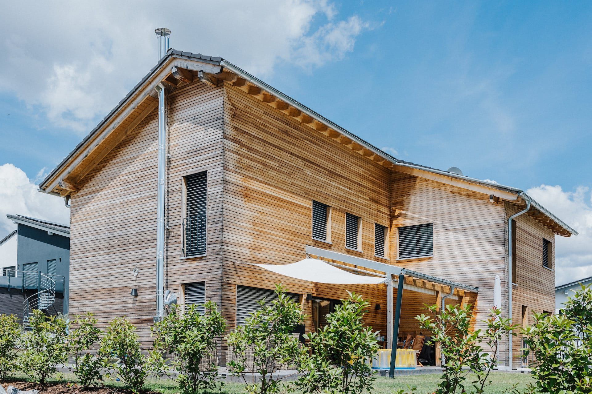 Zweistöckiges Holzhaus mit Satteldach, Fenstern und einer Sitzecke im Freien. Sonniger Tag, blauer Himmel.