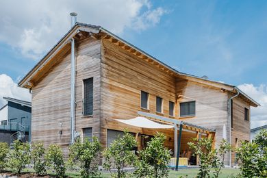 Zweistöckiges Holzhaus mit Satteldach, Fenstern und einer Sitzecke im Freien. Sonniger Tag, blauer Himmel.